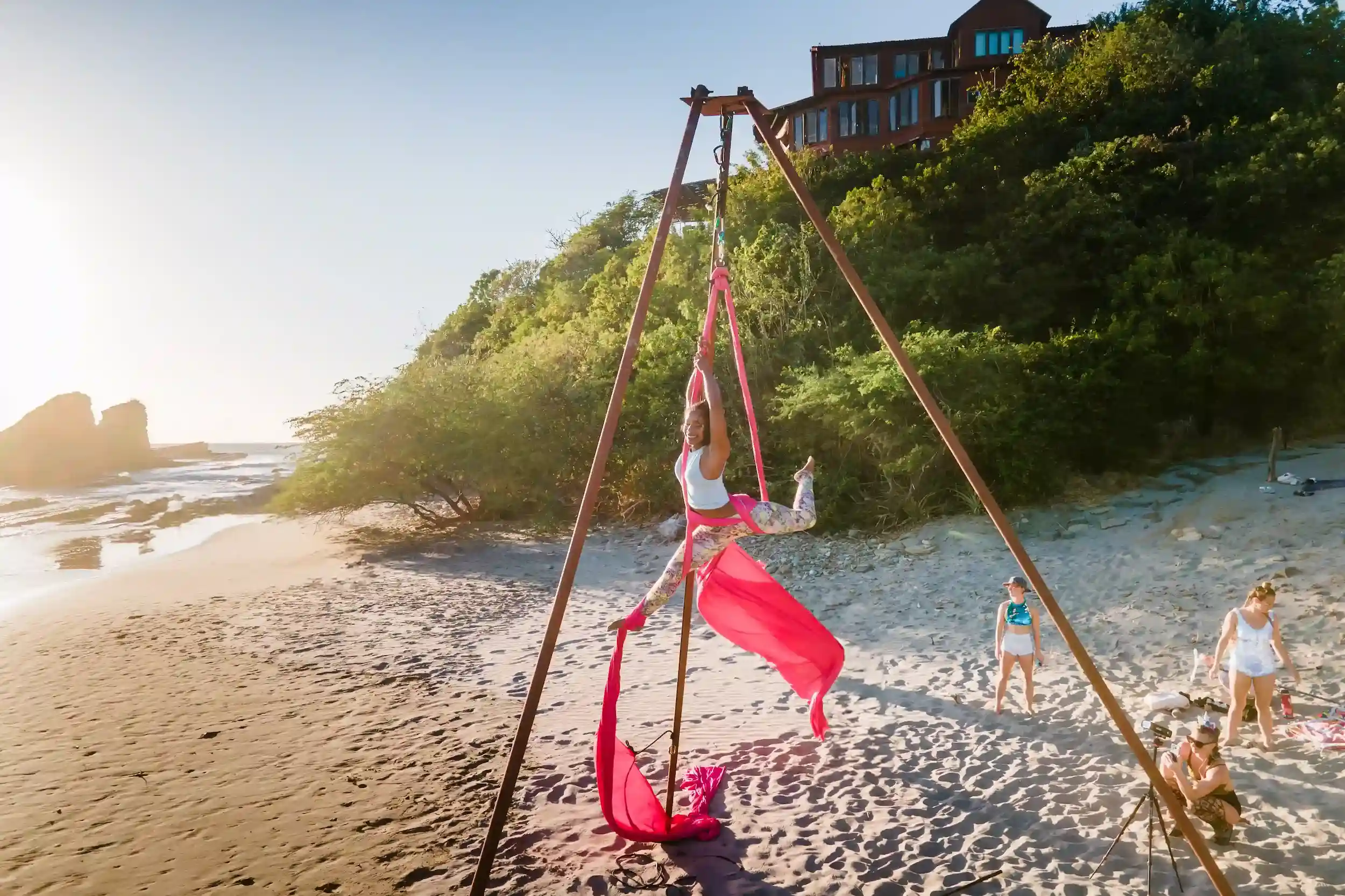 A woman practicing aerial yoga on the beach at sunset while others observe nearby.
