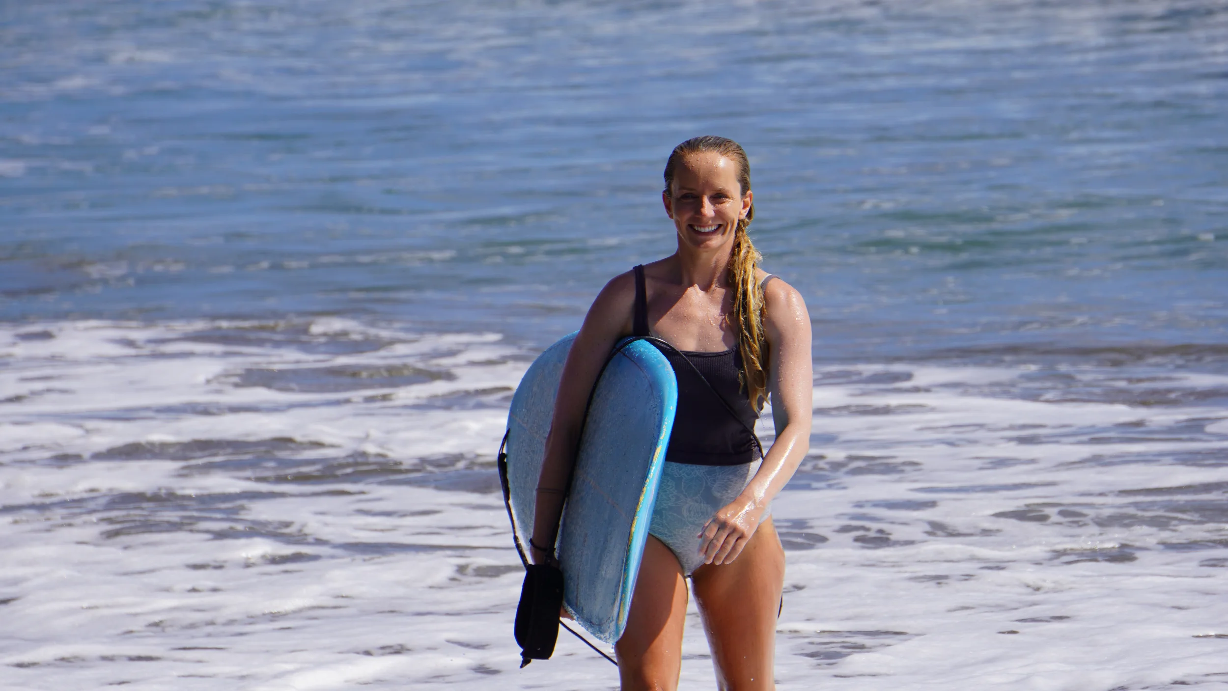 Smiling beginner surfer holding a soft-top surfboard in shallow water at Beginners Bay Popoyo, showing confidence after a surf lesson